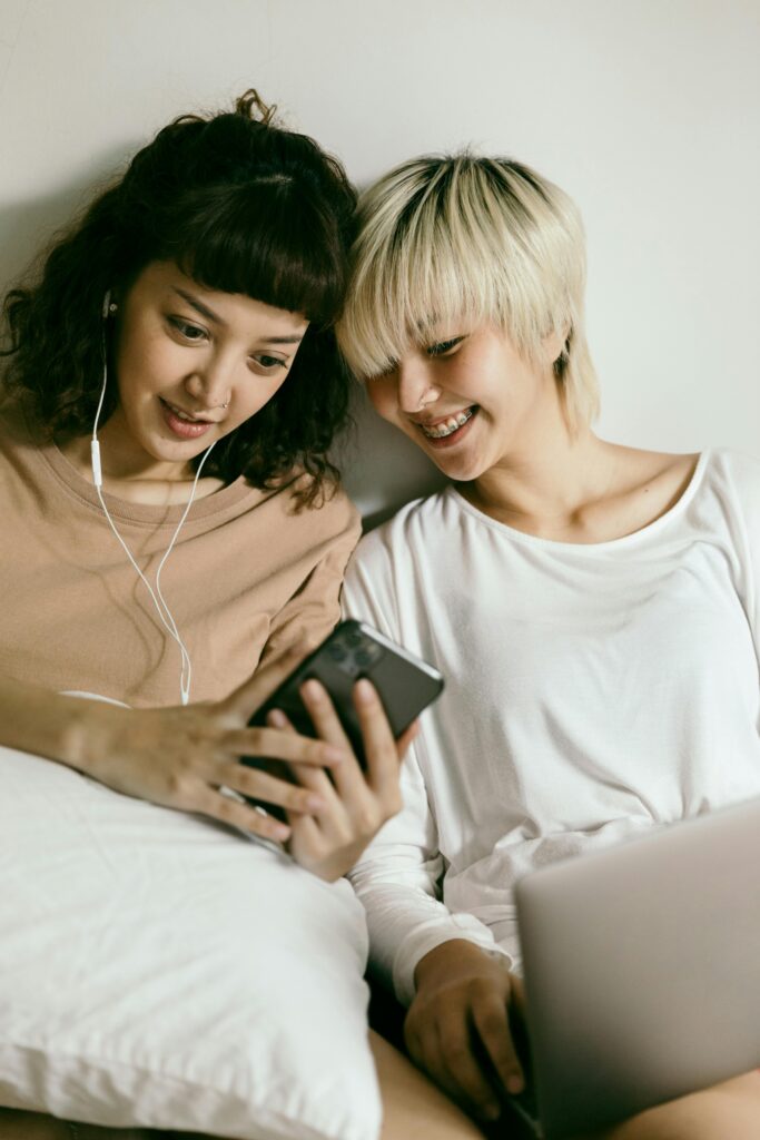 Happy positive young Asian girlfriends in earphones browsing mobile phone and looking at screen while sitting on floor with pillow and netbook together